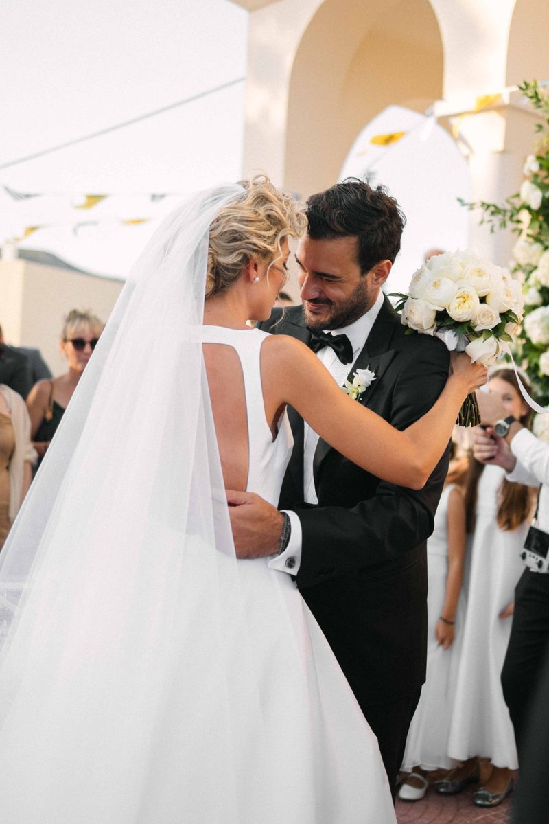 Bride and groom embrace under white arches after the ceremony — wedding photography by John Xenos, Greece.