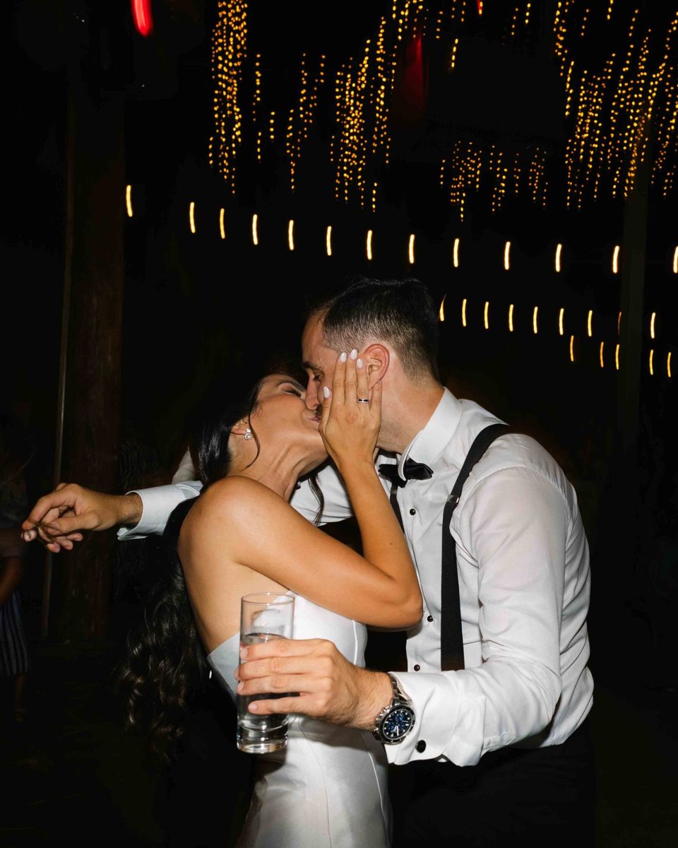 Couple sharing a quiet kiss under warm string lights at the reception — wedding photography by John Xenos, Greece.