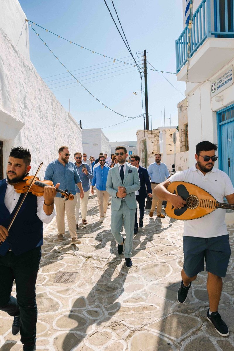 Groom walking through a whitewashed Cycladic alley with traditional musicians — wedding photography by John Xenos, Greece.