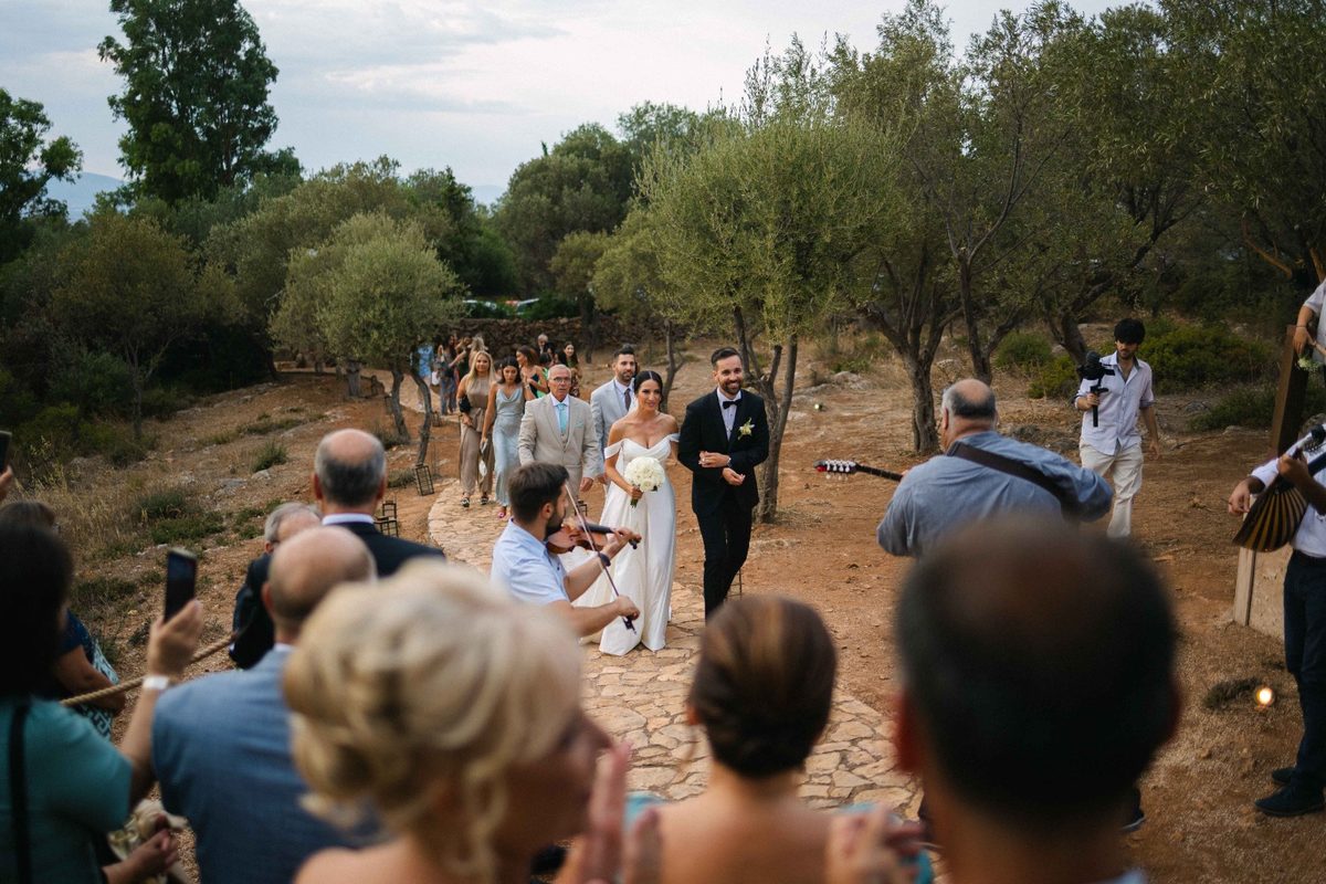 Bridal party walking through an olive grove with live musicians — wedding photography by John Xenos, Greece.